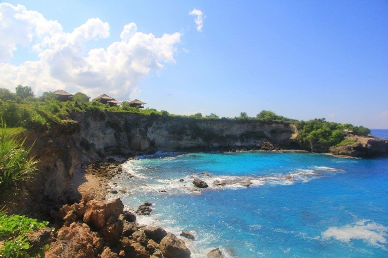 A coastal landscape with clear blue waters, rocky shorelines, and a cliff with vegetation and buildings.
