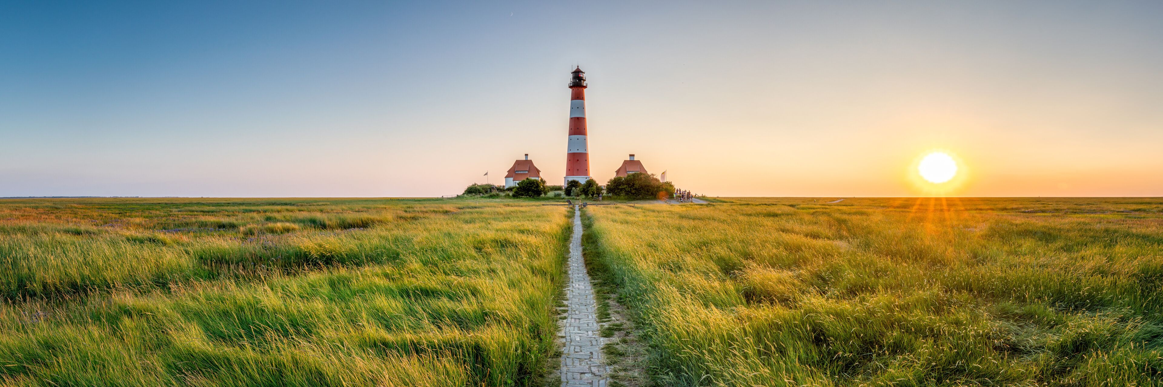 A lighthouse on a grassy field with a path leading to it, and the sun setting in the background.