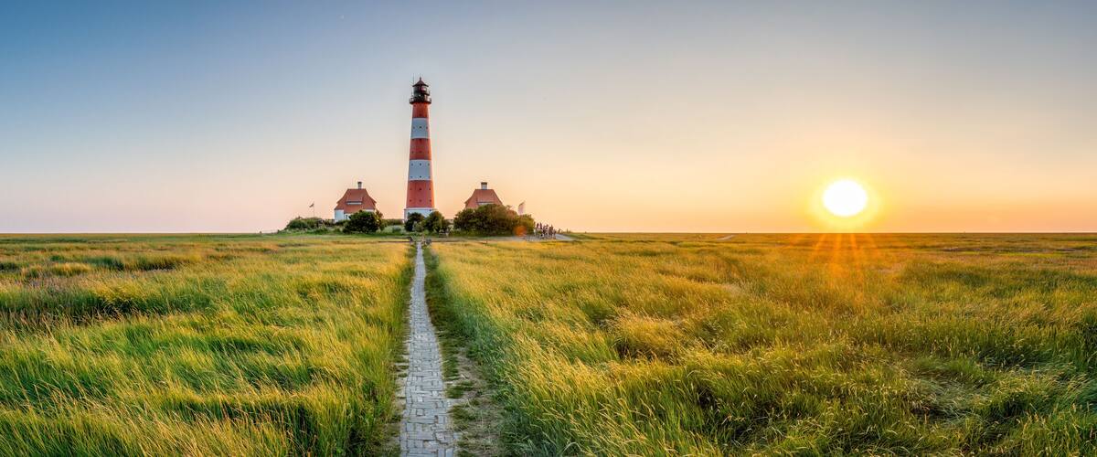 A lighthouse on a grassy field with a path leading to it, and the sun setting in the background.