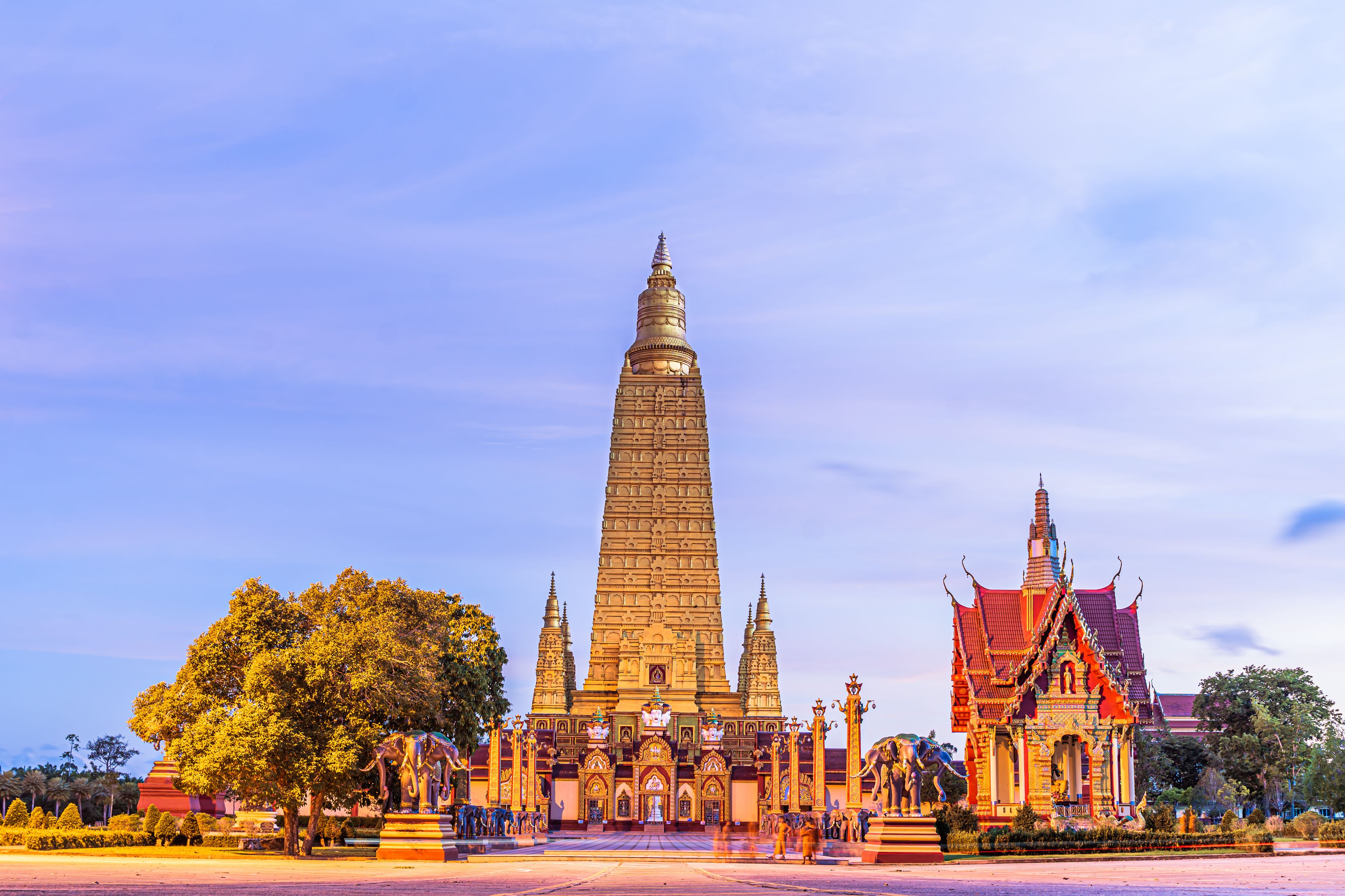 A temple with a tall spire, a smaller adjacent building with a red roof, and a large tree in the foreground.