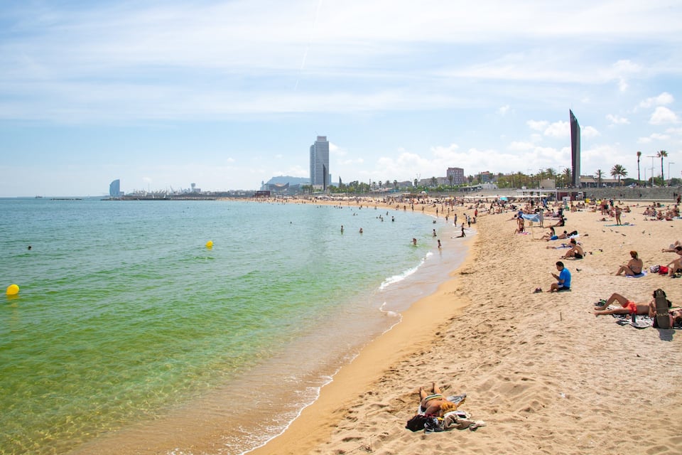 Una spiaggia affollata con acque cristalline, persone che prendono il sole e edifici sullo sfondo.