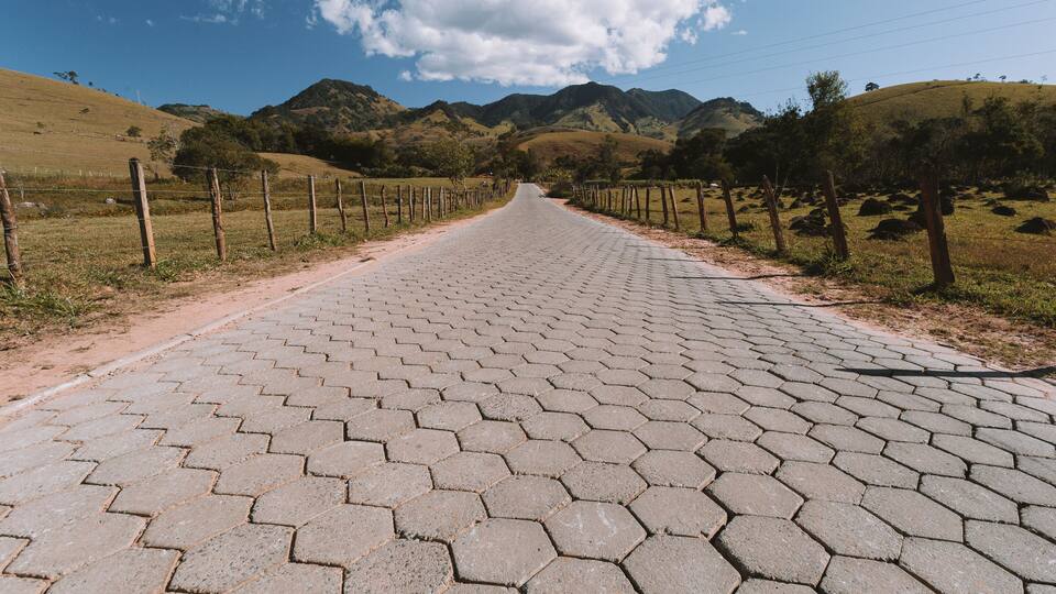 A paved road with hexagonal tiles, flanked by fences and surrounded by rolling hills and mountains.