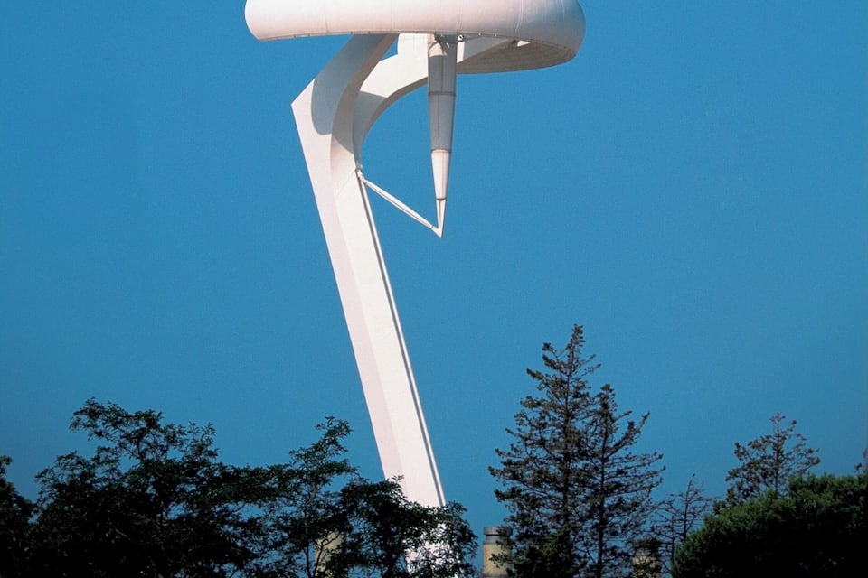 Ein hoher, weißer, moderner Turm mit spitzem Dach vor blauem Himmel.