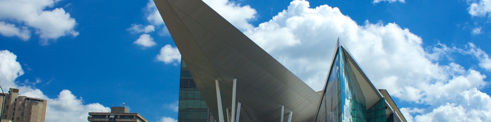 Un edificio moderno con un diseño angular distintivo, rodeado de otras construcciones urbanas y un cielo azul despejado con nubes.