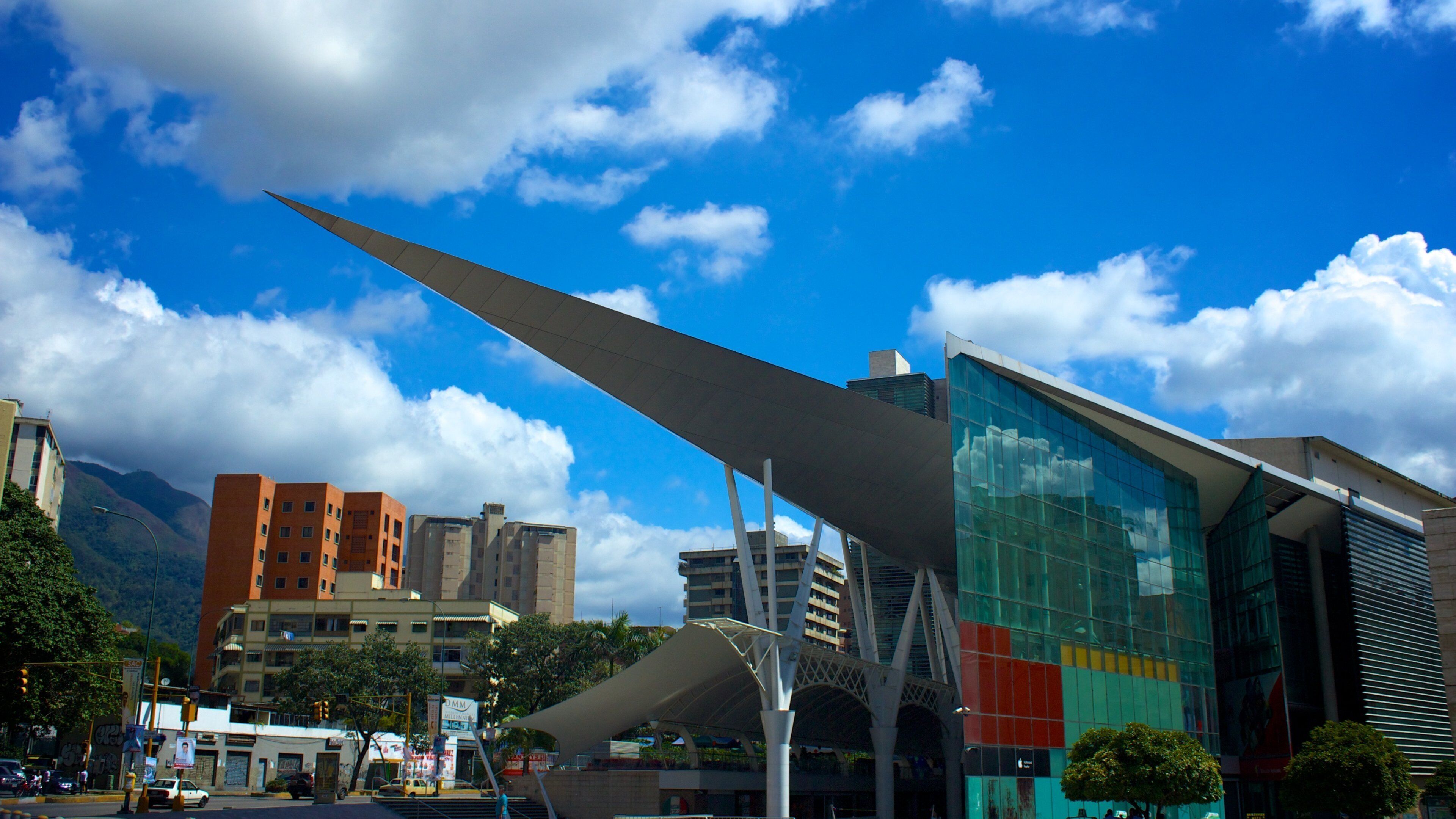 A modern urban landscape with a prominent glass building, a large wing-like structure, and a mountain in the background.