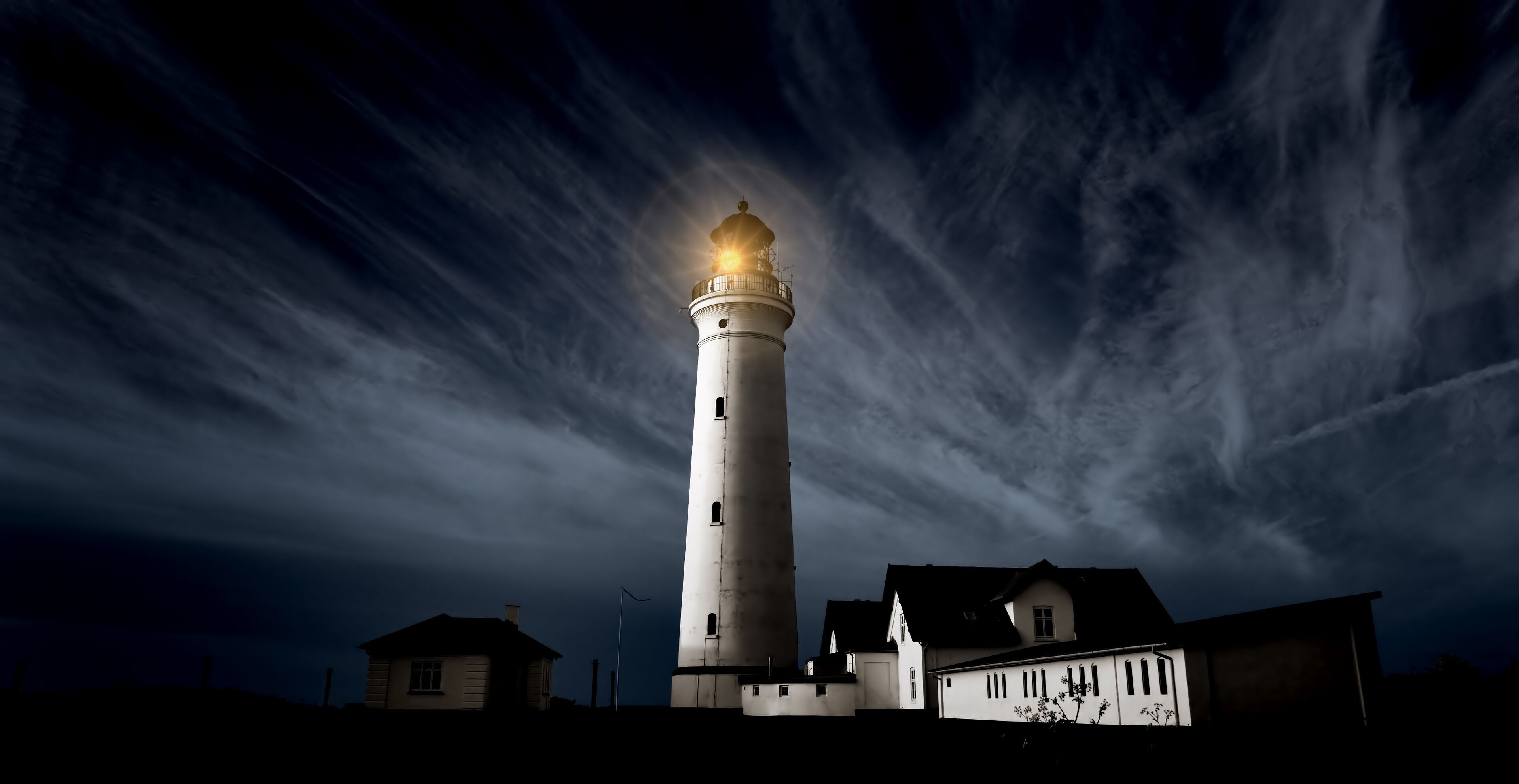 A lighthouse with a bright light at the top, surrounded by houses and a cloudy sky.