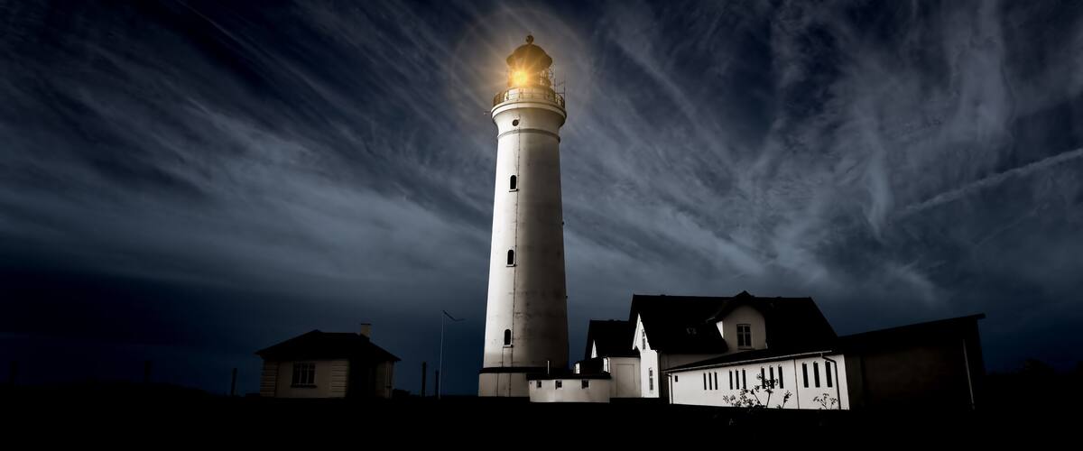 A lighthouse with a bright light at the top, surrounded by houses and a cloudy sky.