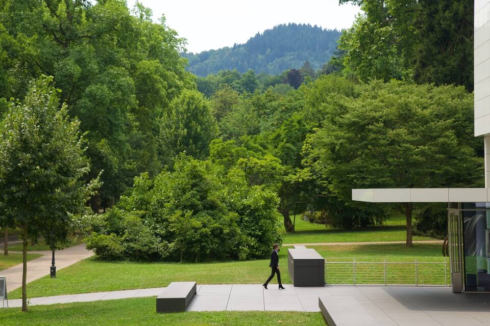Un edificio moderno con ingresso in vetro, immerso nel verde tra alberi rigogliosi e un'area erbosa.
