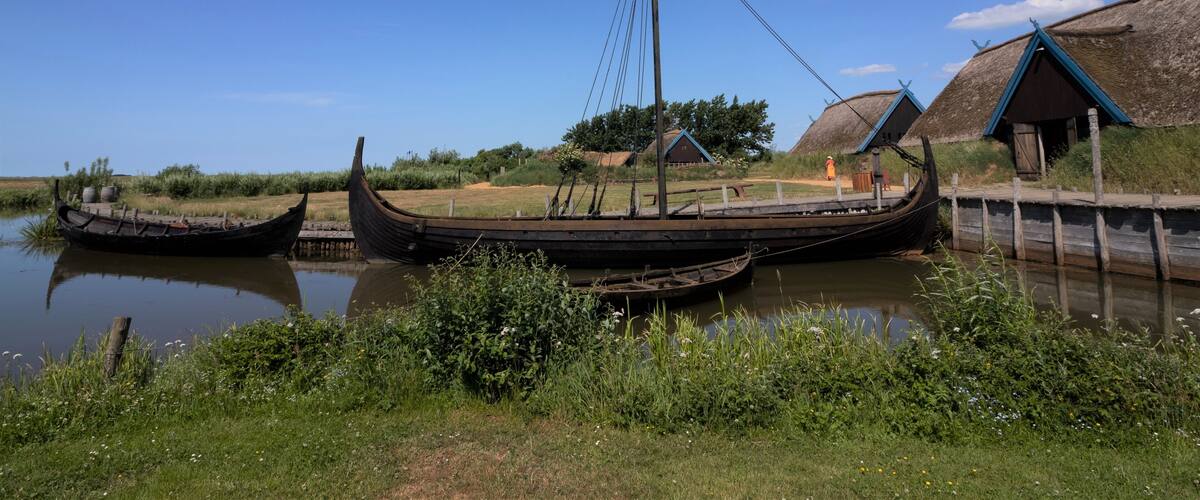 A traditional boat docked by a riverbank with thatched-roof buildings in the background.