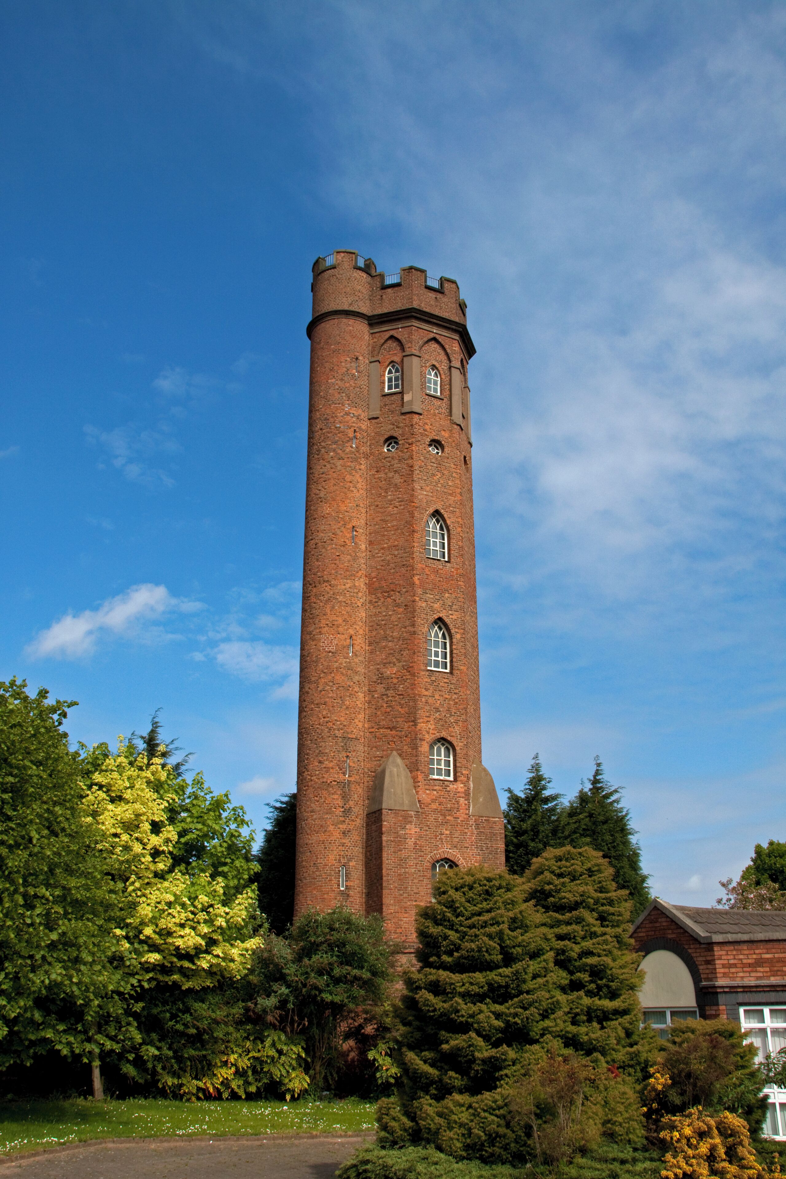 A tall brick tower with arched windows and a crenellated top, surrounded by lush greenery and trees.