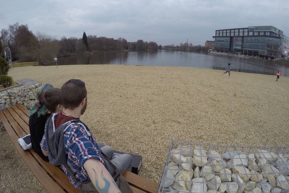A lakeside park with a wooden bench, a metal fence, and a modern building in the background.