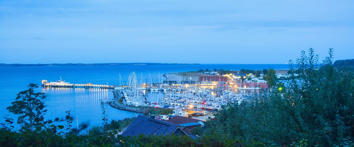 A marina at dusk with boats docked, a pier extending into the water, and buildings in the background.
