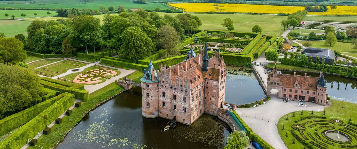 A historic castle with a moat, surrounded by manicured gardens and fields.