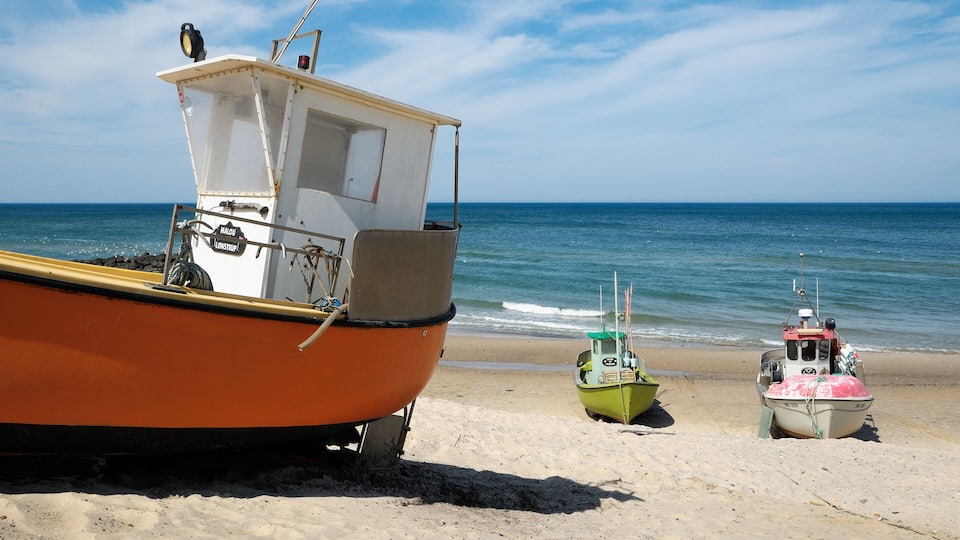 A beach with three boats: an orange boat with a cabin, a green boat, and a pink boat.