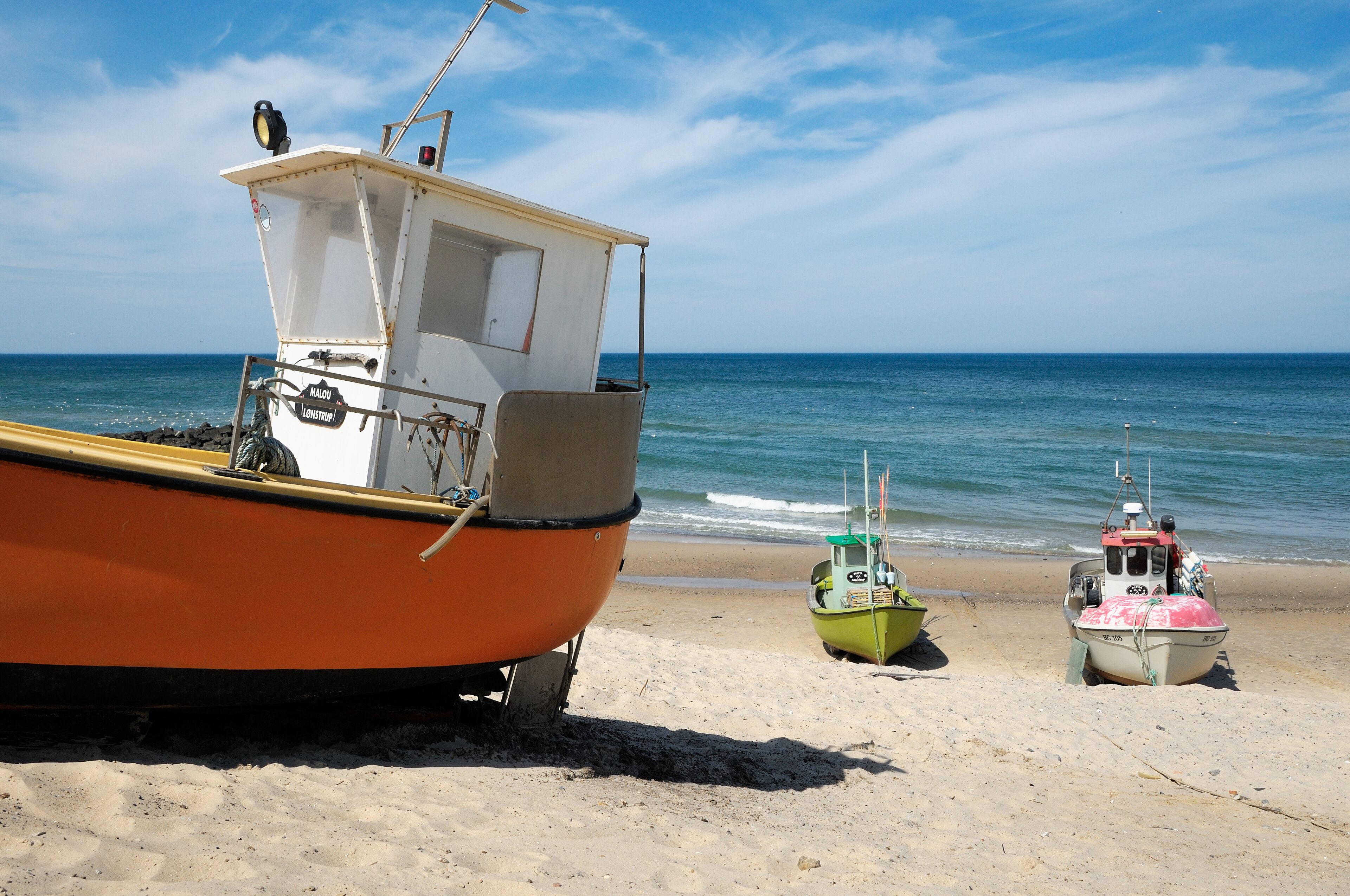 A beach with three boats: an orange boat with a cabin, a green boat, and a pink boat.
