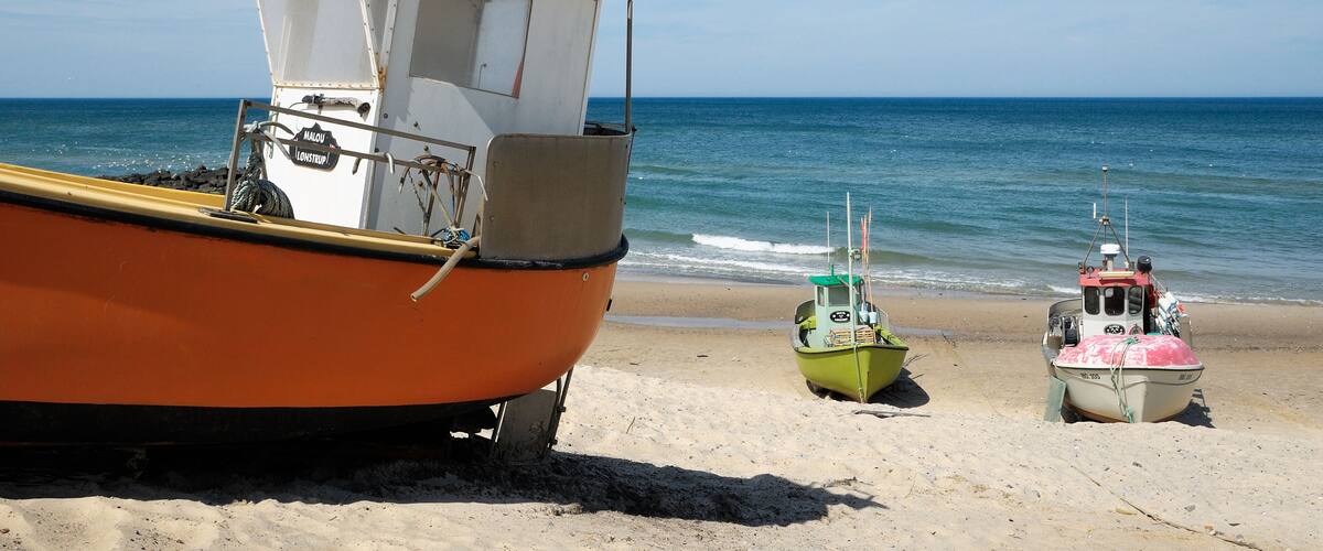 A beach with three boats: an orange boat with a cabin, a green boat, and a pink boat.