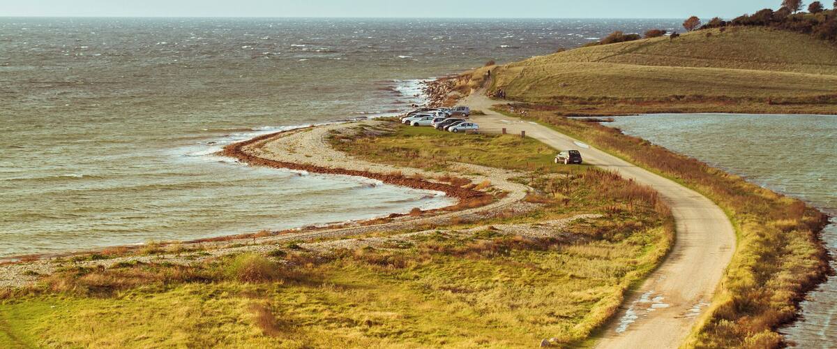 A coastal road winding along a grassy embankment with parked cars and a lighthouse in the distance.
