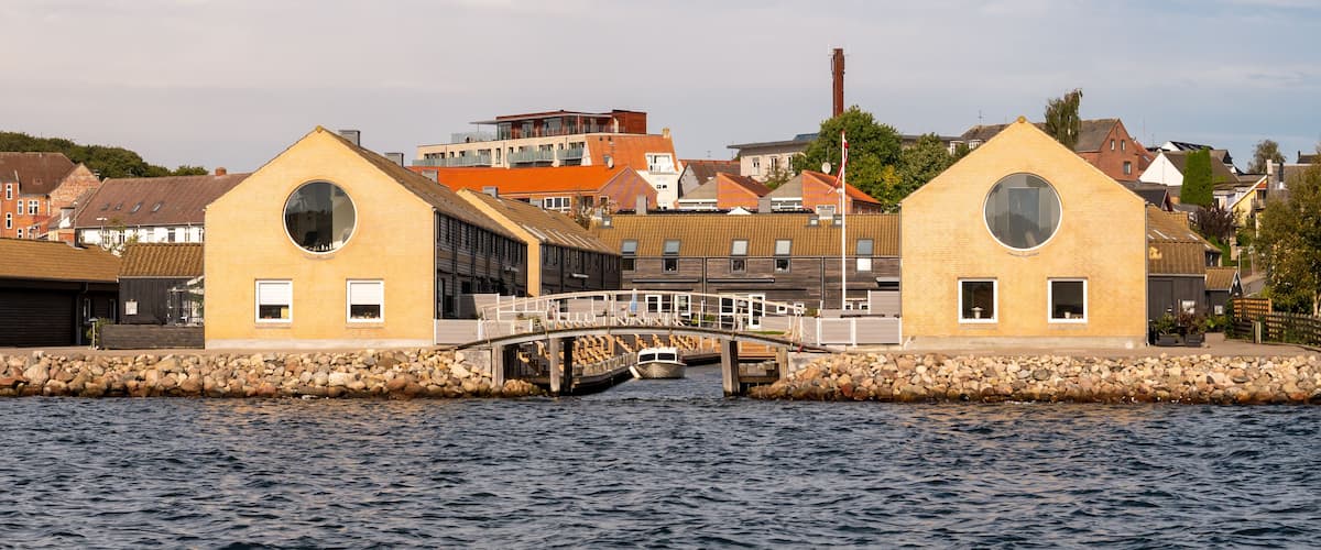 A waterfront with two yellow buildings with circular windows, a bridge, and a boat in the water.