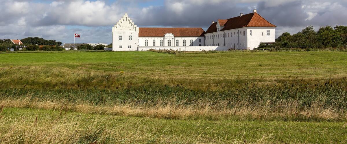 A large white building with a red roof in a grassy field under a partly cloudy sky.