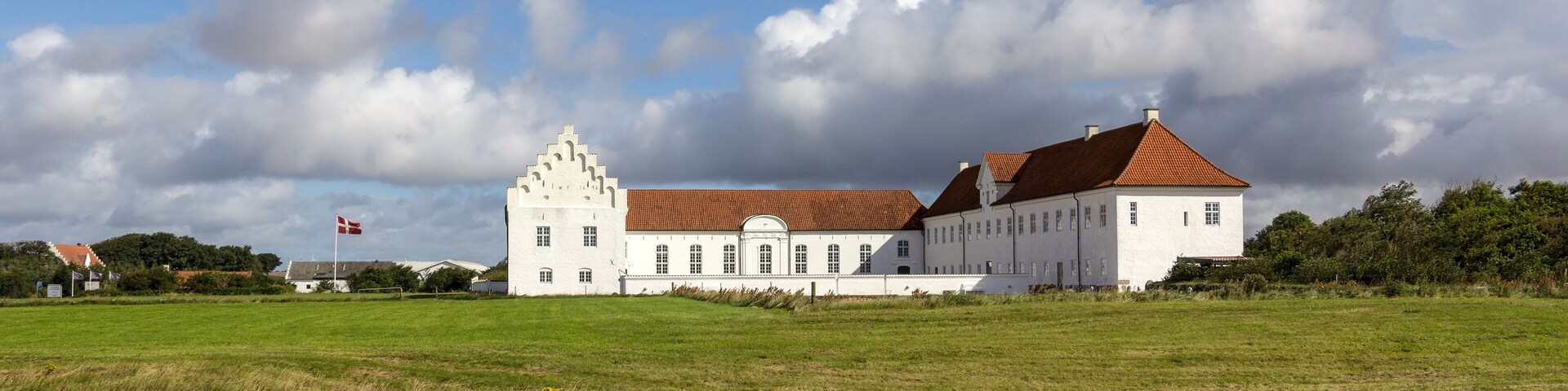 A large white building with a red roof in a grassy field under a partly cloudy sky.