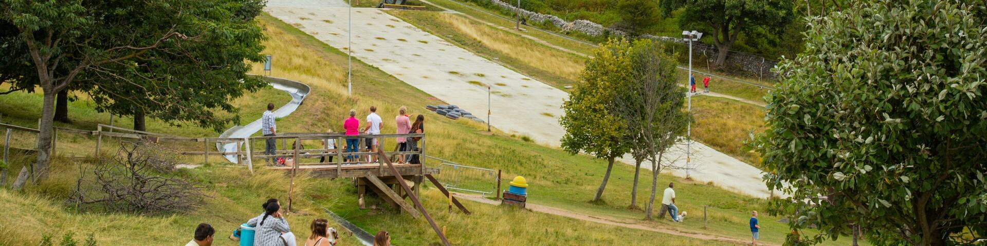 A park with a metal slide, grassy areas, trees, and a rocky hillside in the background.
