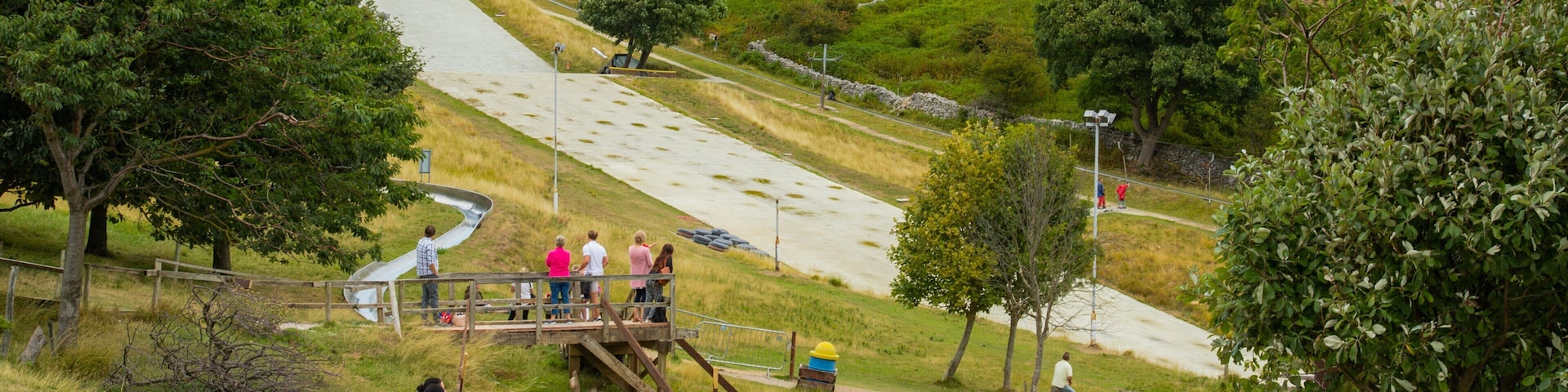 Um parque com um escorrega de metal, áreas gramadas, árvores e uma encosta rochosa ao fundo.