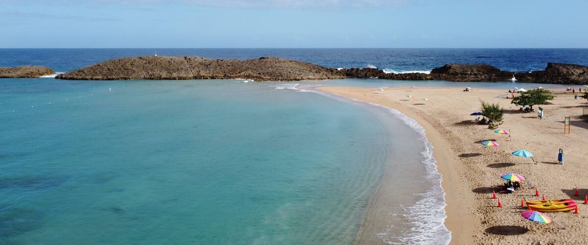 A beach with clear turquoise waters, a rocky outcrop, and colorful umbrellas.