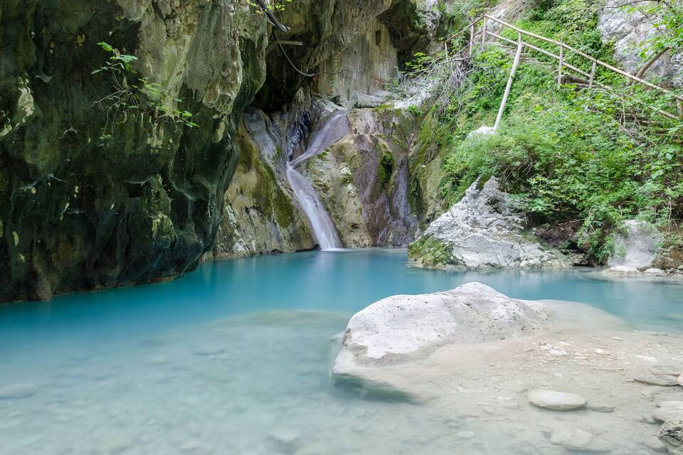 En turkis pool med et vandfald, omgivet af klippepartier og frodig vegetation.