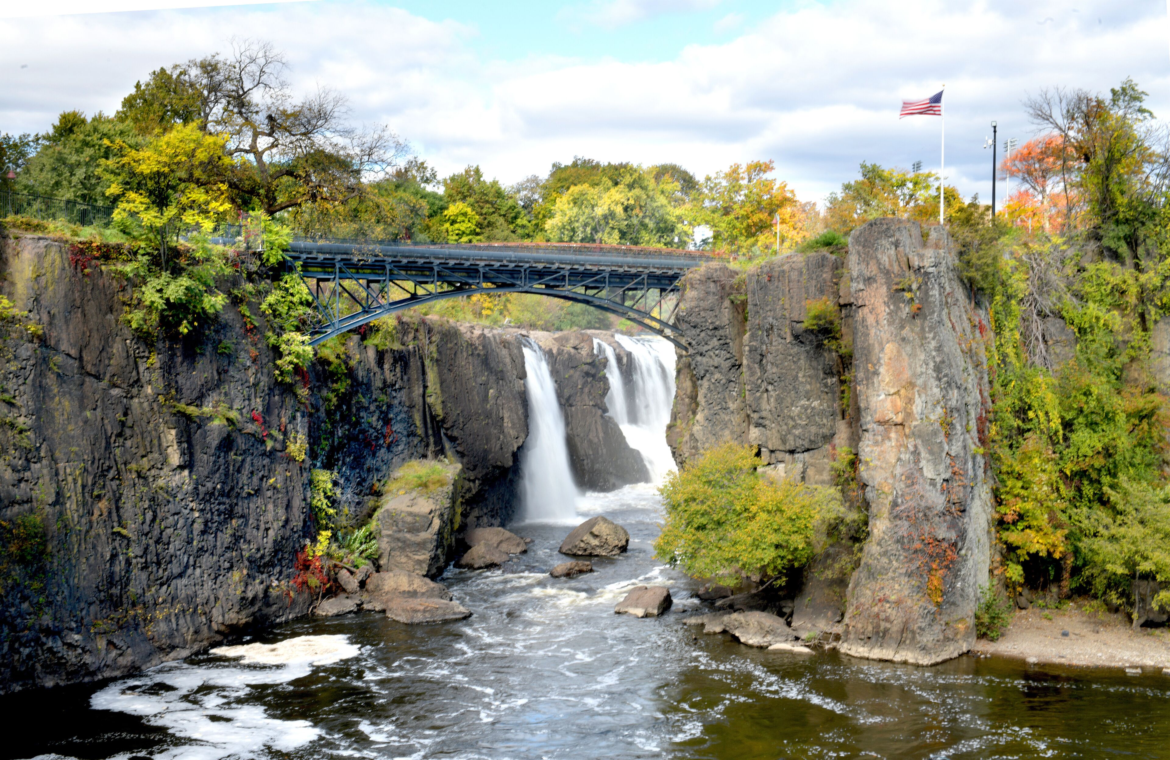 A waterfall under a bridge with an American flag, surrounded by autumn foliage and rocky terrain.
