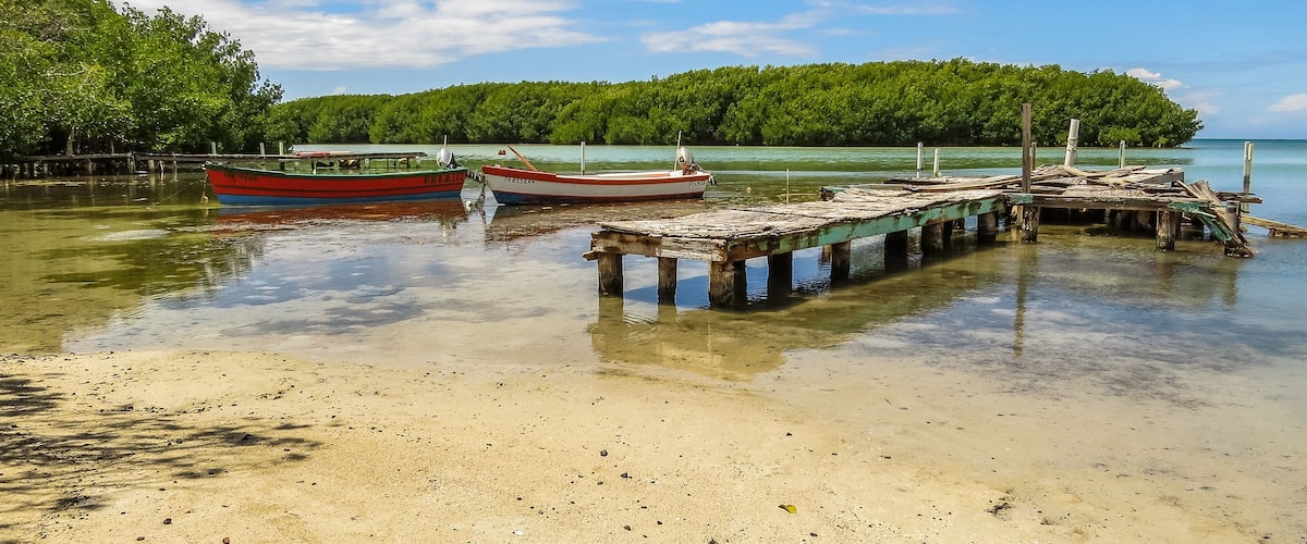 Dos barcos amarrados a un muelle de madera, con una playa arenosa y una isla boscosa al fondo.