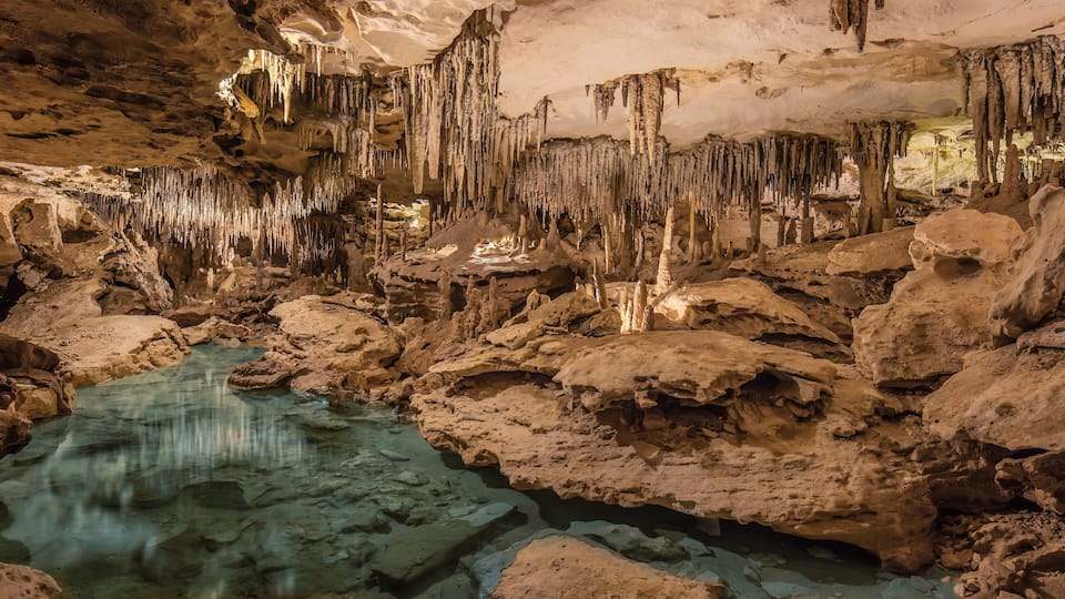 A cave with stalactites and stalagmites, a water pool, and rocky formations.