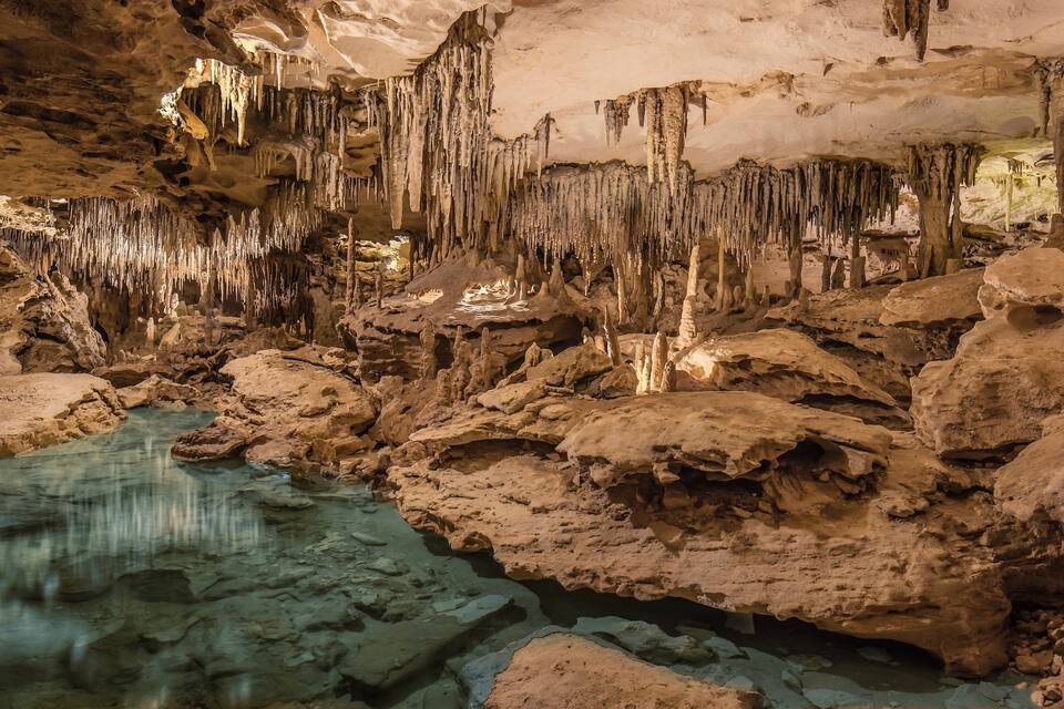 A cave with stalactites and stalagmites, a water pool, and rocky formations.