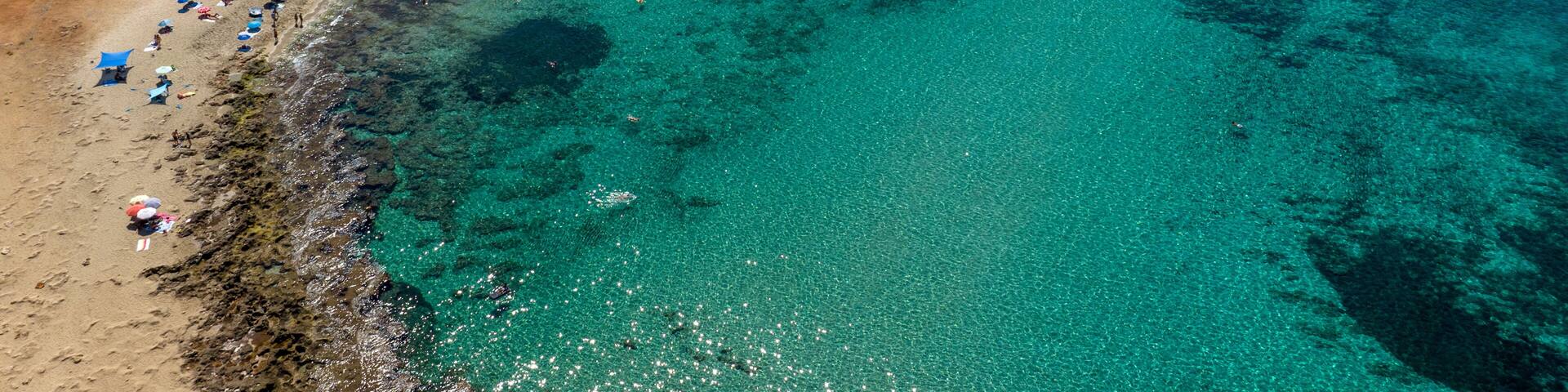 An aerial view of a beach with clear turquoise waters, a long stretch of umbrellas, and a rocky coastline.