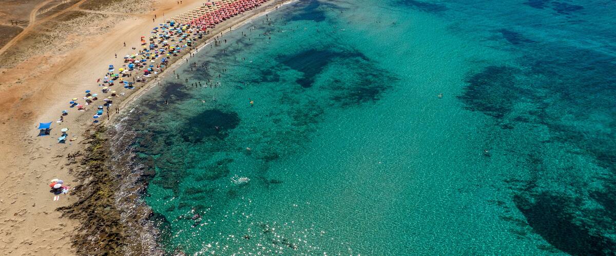 An aerial view of a beach with clear turquoise waters, a long stretch of umbrellas, and a rocky coastline.