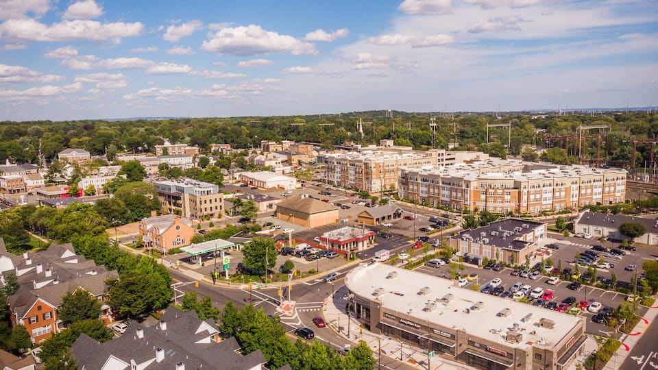 A cityscape with a mix of residential and commercial buildings, a large parking lot, and a clear blue sky with scattered clouds.