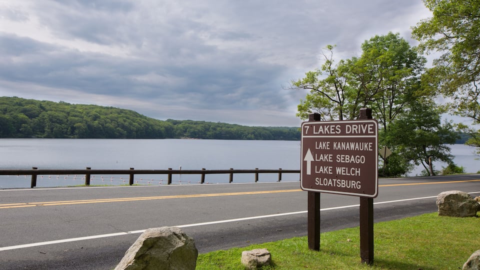 A road sign by a lake with directions to 7 Lakes Drive, Lake Kanawauke, Lake Sebago, Lake Welch, and Sloatsburg.