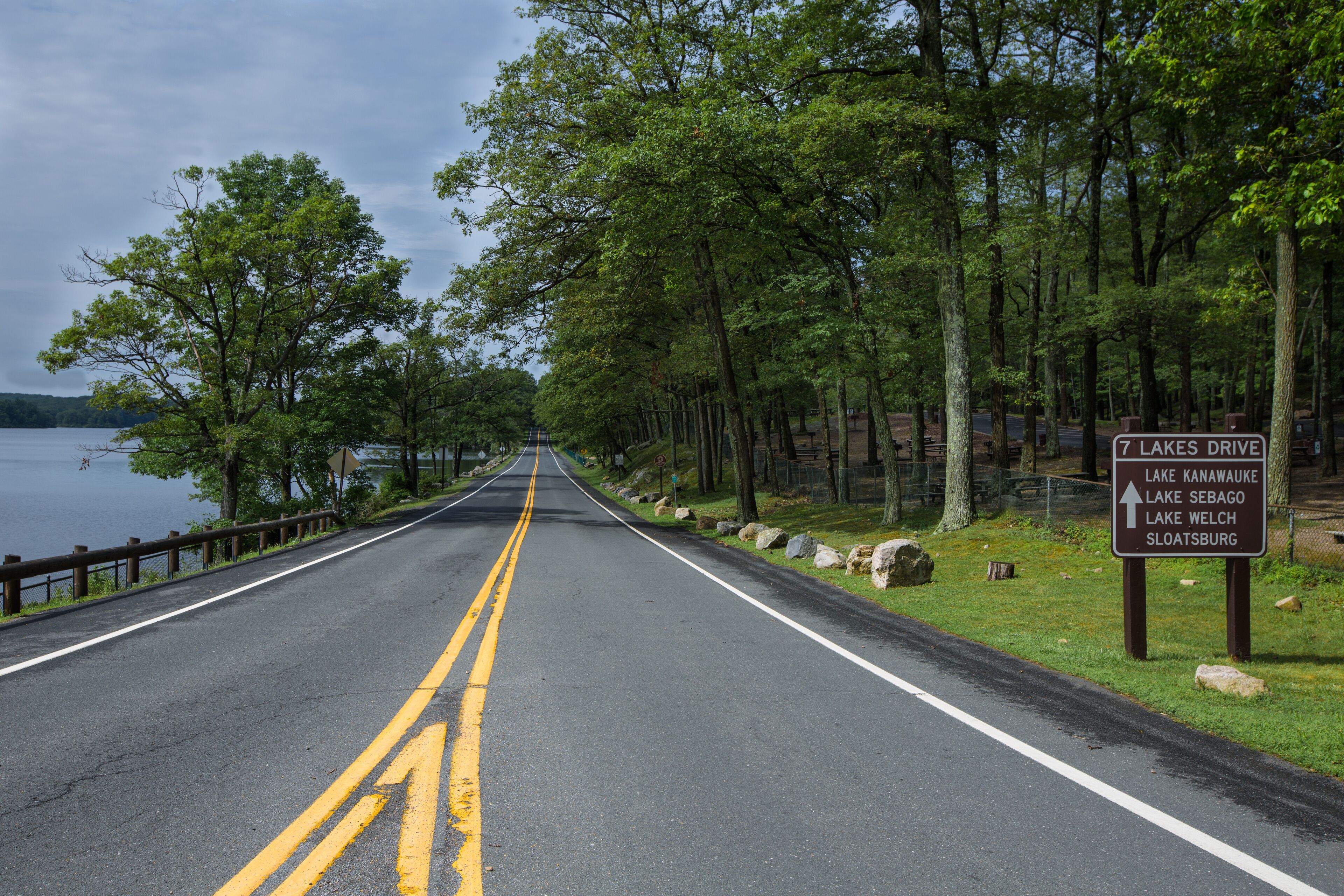 A road with a sign indicating directions to 7 Lakes Drive, Lake Kanawauke, Lake Sebago, Lake Welch, and Sloatsburg.