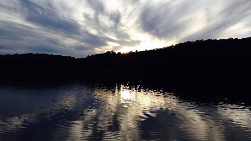 A calm lake with reflections of clouds and a silhouette of trees against a sunset sky.
