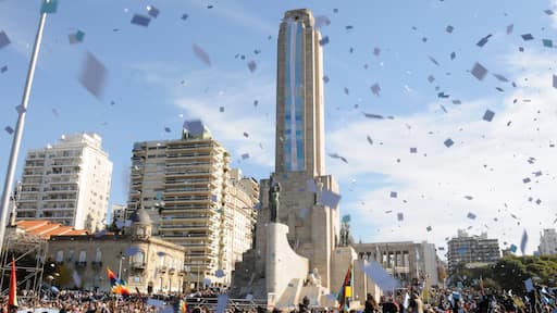 A large monument with a tall obelisk, surrounded by modern buildings and a crowd of people.