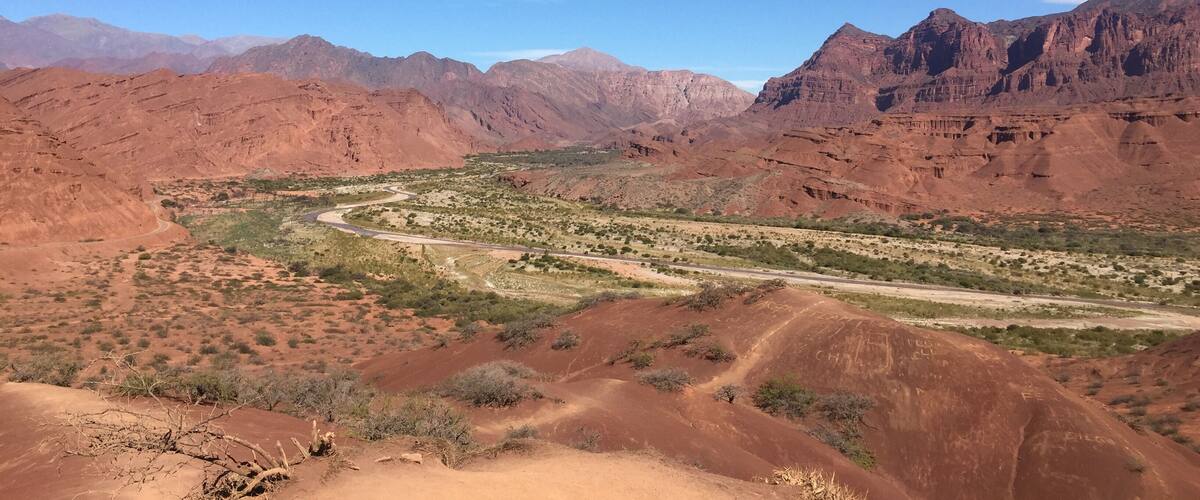 A desert landscape with red rock formations, a winding road, and distant mountains under a clear blue sky.