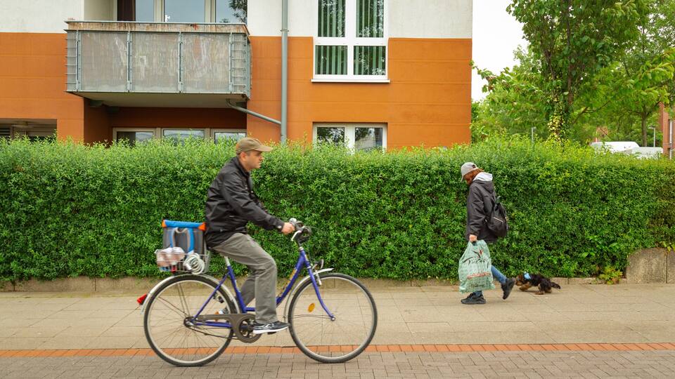 En man som cyklar på en asfalterad stig, en kvinna som promenerar med en hund och en byggnad med orange och vita väggar, med en grön häck i bakgrunden.