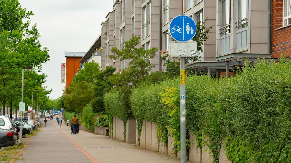 Un trottoir en ville, avec des voitures garées, un panneau indiquant une piste cyclable, et une personne qui marche.