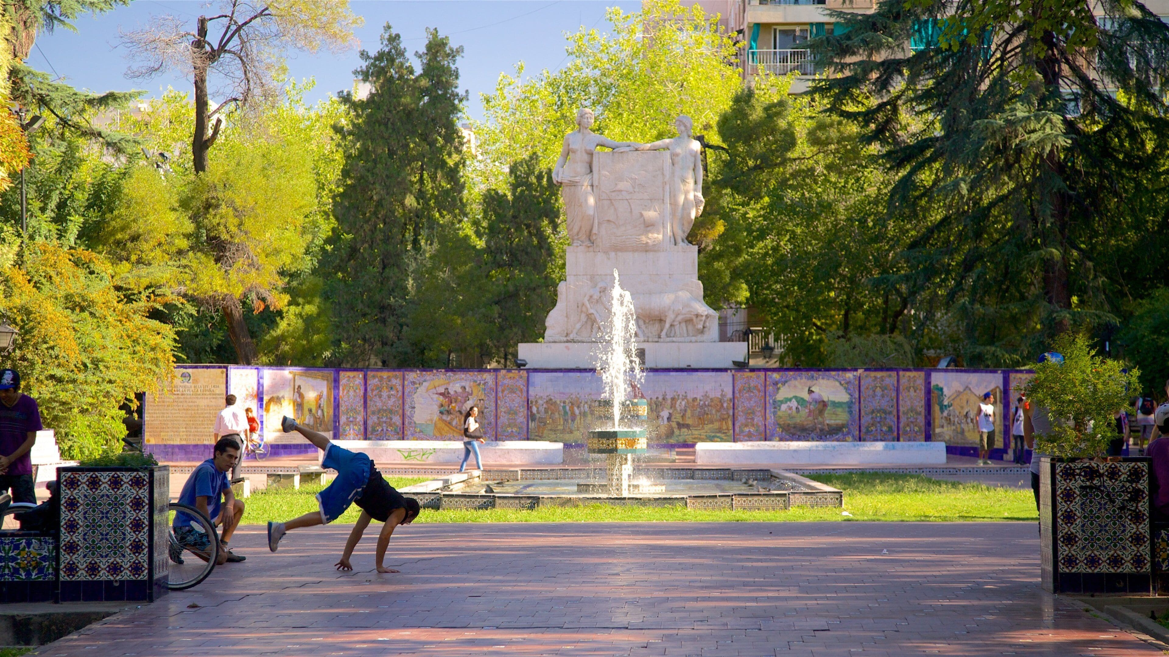 Um parque com uma fonte, uma estátua e um muro de azulejos com murais.