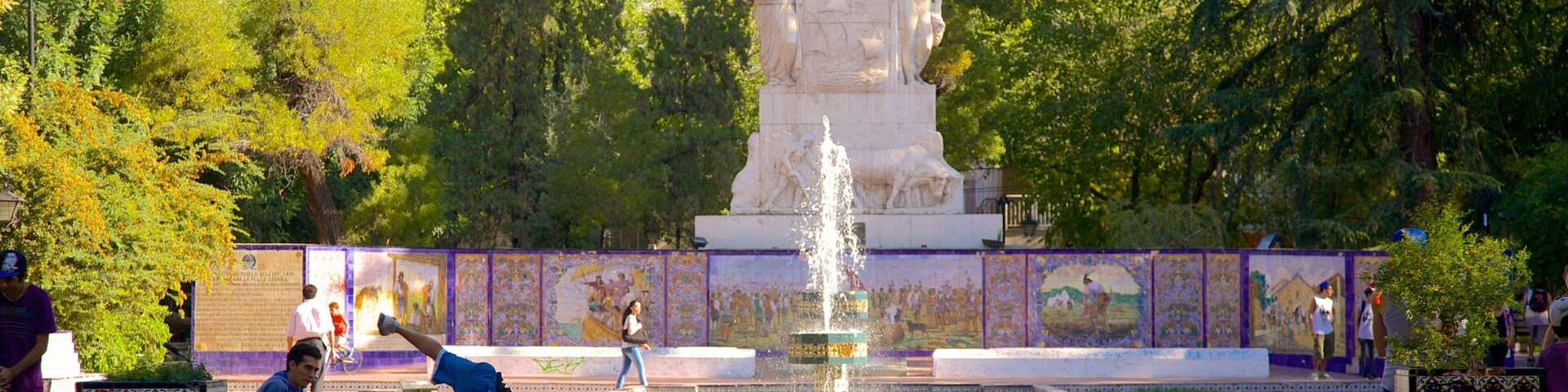 Um parque com uma fonte, uma estátua e um muro de azulejos com murais.