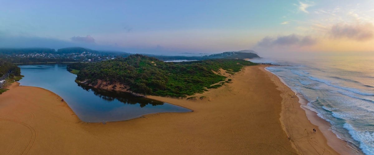 A coastal landscape with a sandy beach, a calm lake, and a forested hill.