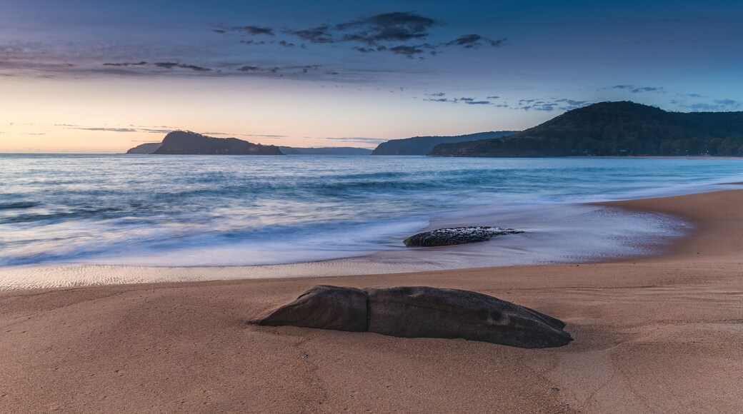 A beach with a large rock in the foreground, calm waves, and distant hills under a twilight sky.