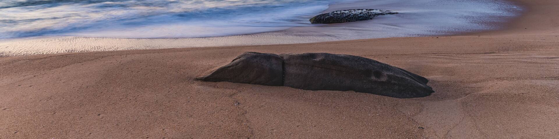 A beach with a large rock in the foreground, calm waves, and distant hills under a twilight sky.