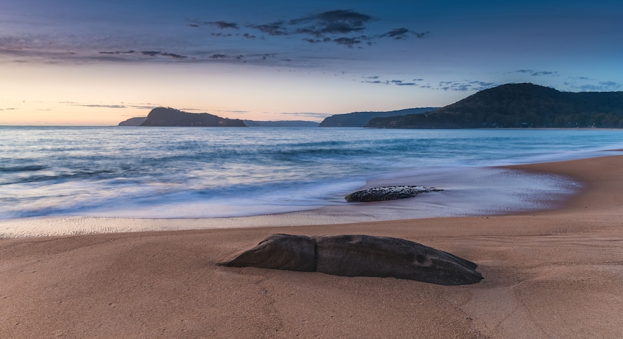 Una playa con una roca grande al frente, olas tranquilas y colinas distantes bajo un cielo crepuscular.