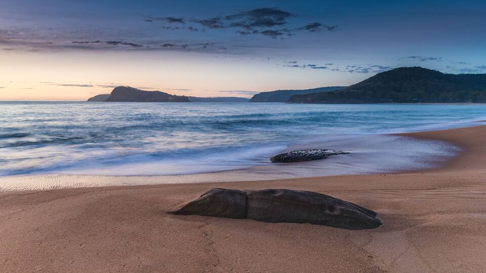 A beach with a large rock in the foreground, calm waves, and distant hills under a twilight sky.