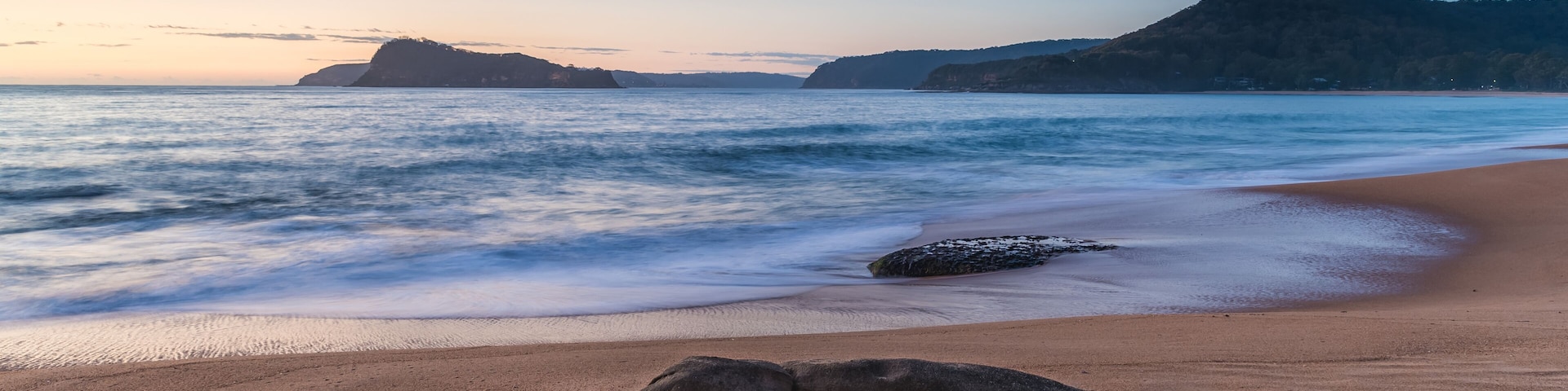 A beach with a large rock in the foreground, calm waves, and distant hills under a twilight sky.